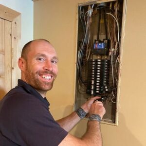 A man smiles while working on wiring inside an open electrical panel mounted on a wall.