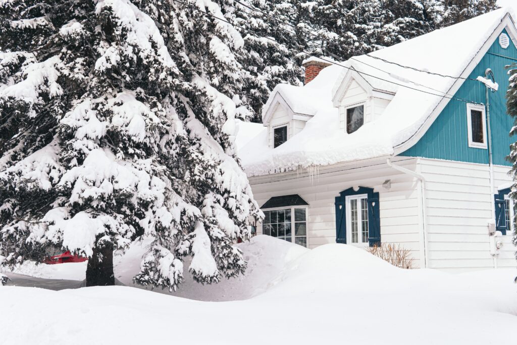 Small white-and-teal Cape-style home with large pine tree in front, all covered in feet of snow.