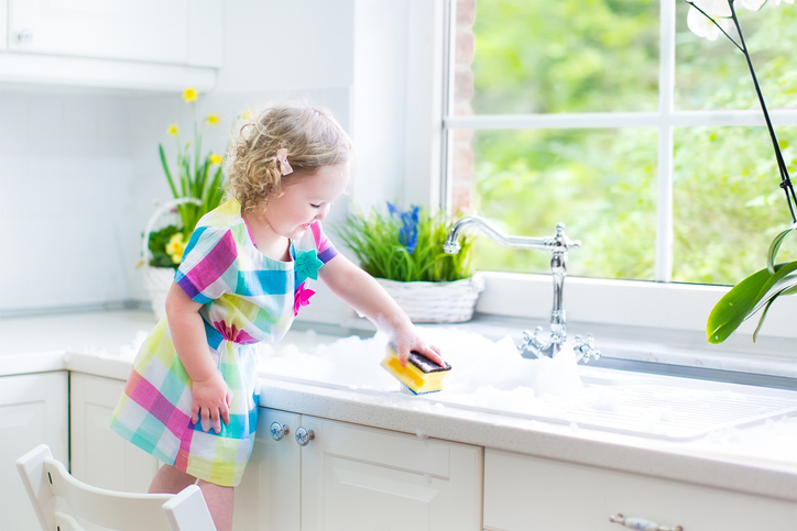 Cute curly toddler girl washing dishes, cleaning with sponge