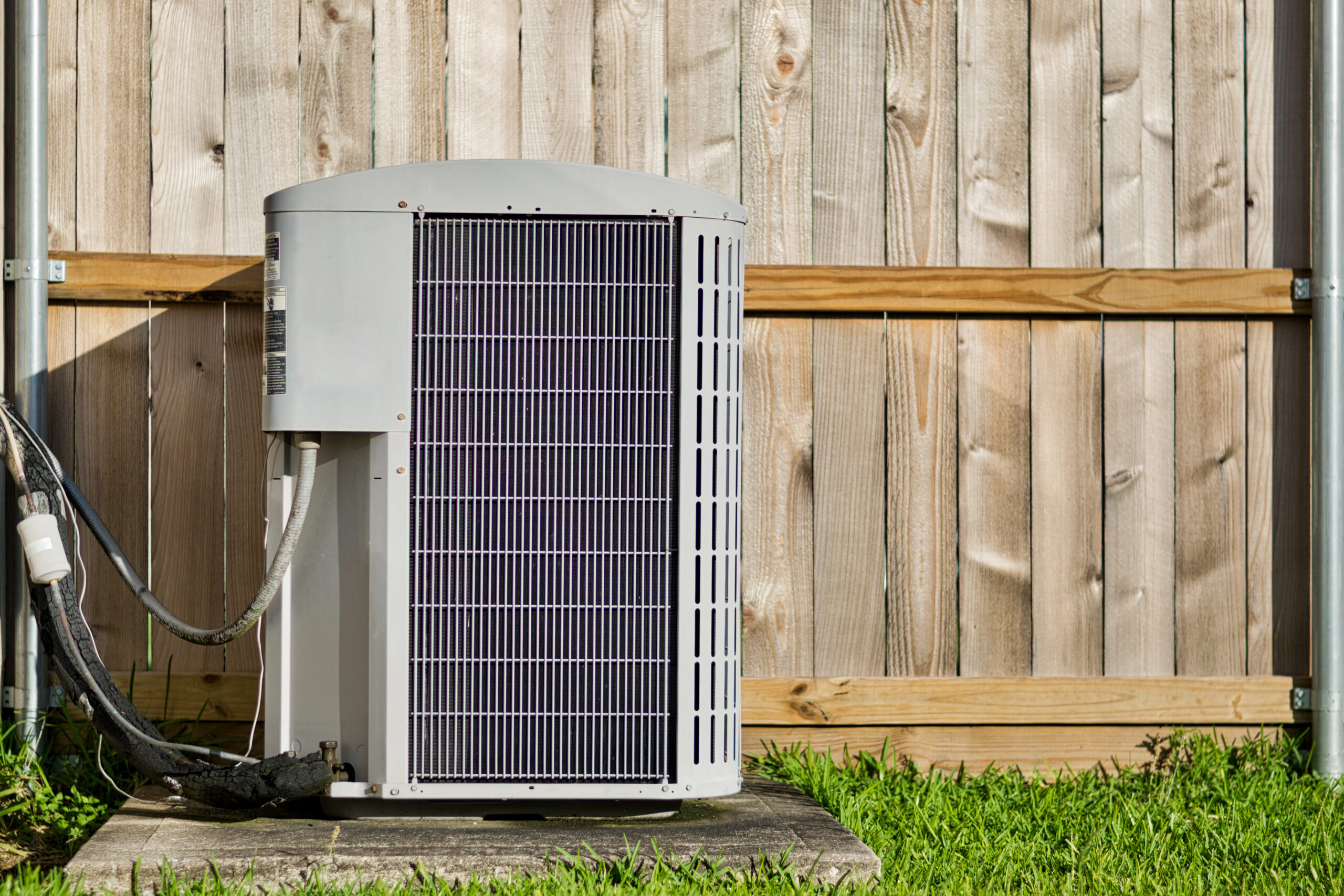 Central air conditioning unit in a residential backyard in front of a wooden fence and lawn area